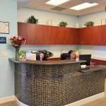 A curved, dark-tiled reception desk with a granite countertop, situated in front of wood-grain cabinetry and a bright office space