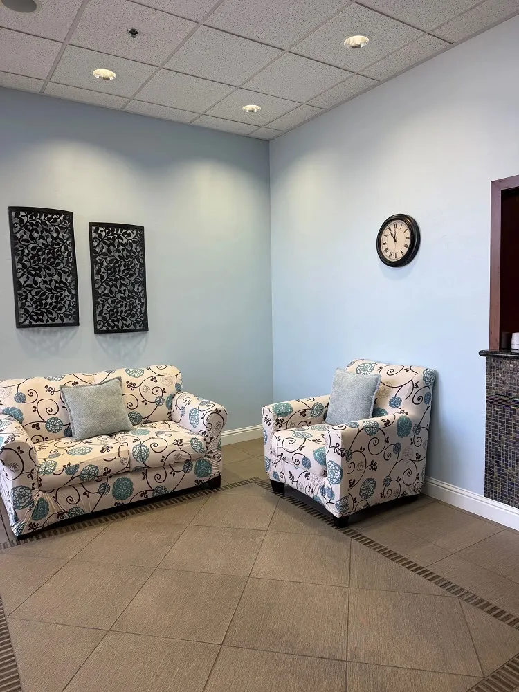 A bright waiting room with light blue walls, tiled floors, and two white sofas with a blue and pink floral pattern.